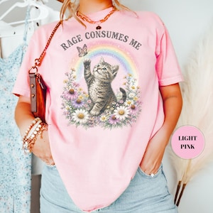 May include: Light pink t-shirt with a graphic of a kitten reaching for a butterfly in front of a rainbow. The text "Rage Consumes Me" is above the graphic. The shirt is a casual style.