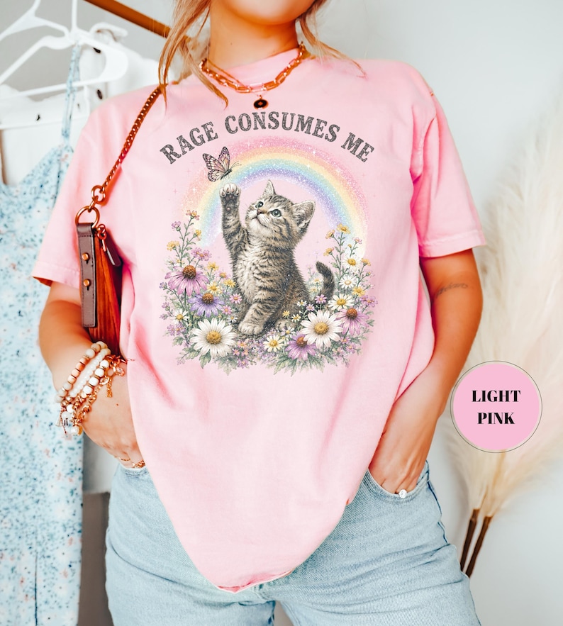 May include: Light pink t-shirt with a graphic of a kitten reaching for a butterfly in front of a rainbow. The text "Rage Consumes Me" is above the graphic. The shirt is a casual style.