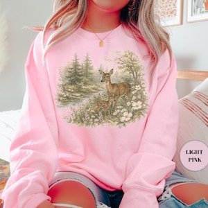 May include: Light pink sweatshirt featuring a detailed illustration of two deer in a forest setting. The artwork depicts a doe and a fawn amidst trees, flowers, and a small stream. The sweatshirt is a solid light pink color.