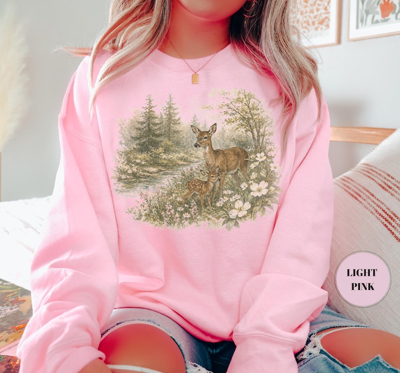 May include: Light pink sweatshirt featuring a detailed illustration of two deer in a forest setting. The artwork depicts a doe and a fawn amidst trees, flowers, and a small stream. The sweatshirt is a solid light pink color.