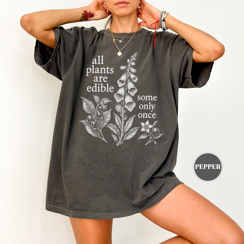 以下が含まれることがあります： チャコールグレーの半袖Tシャツ。ジギタリスの植物とベリーの白いボタニカルイラストが描かれています。「all plants are edible some only once」というテキストがプリントされています。クルーネックでゆったりとしたフィット感のヴィンテージ風Tシャツです。