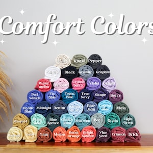 May include: A display of rolled-up t-shirts in various colors, arranged in a pyramid shape. The image features the text "Comfort Colors" at the top, with color names like "Black," "Blue," and "Berry" labeling each shirt. The shirts are neatly stacked.