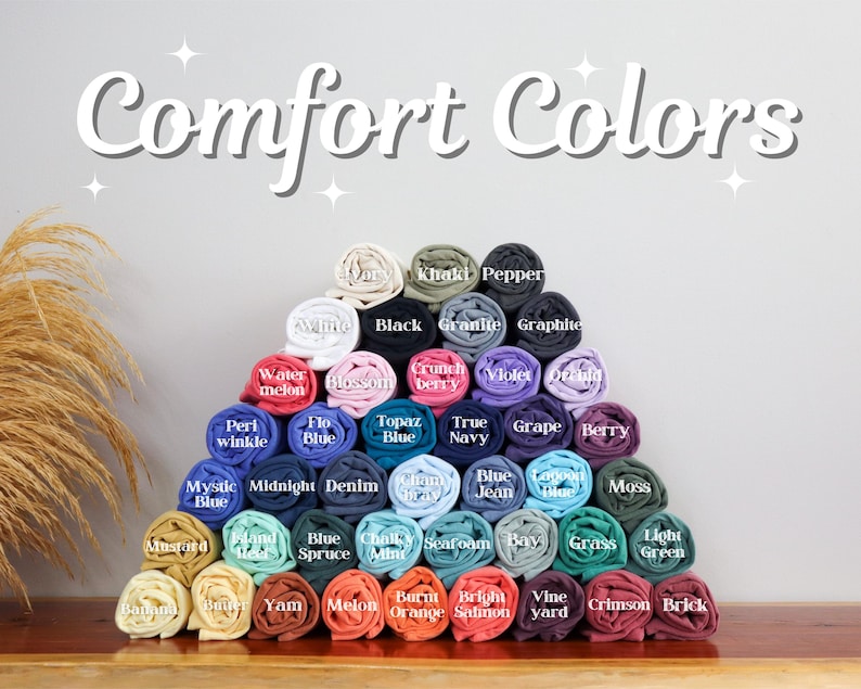 May include: A display of rolled-up t-shirts in various colors, arranged in a pyramid shape. The image features the text "Comfort Colors" at the top, with color names like "Black," "Blue," and "Berry" labeling each shirt. The shirts are neatly stacked.