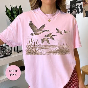 Peut inclure: Un t-shirt rose clair &agrave; manches courtes avec une impression graphique repr&eacute;sentant un vol d'oies au-dessus d'un marais avec des roseaux. L'illustration est rendue dans des tons de brun et de beige, montrant les oiseaux en plein vol.