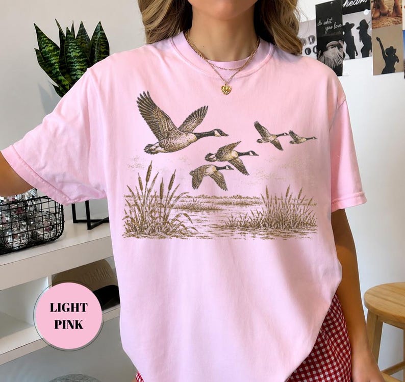 Peut inclure: Un t-shirt rose clair &agrave; manches courtes avec une impression graphique repr&eacute;sentant un vol d'oies au-dessus d'un marais avec des roseaux. L'illustration est rendue dans des tons de brun et de beige, montrant les oiseaux en plein vol.