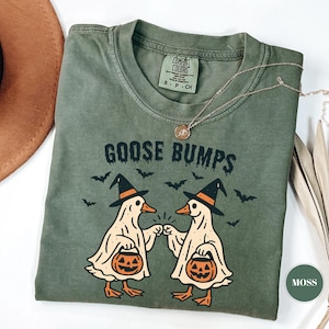 May include: Moss green t-shirt with the text "GOOSE BUMPS" and a graphic of two geese dressed as ghosts, wearing witch hats and holding pumpkin baskets. Bats are flying above the geese. A gold necklace is draped across the shirt.