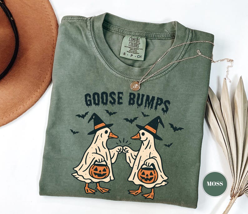 May include: Moss green t-shirt with the text "GOOSE BUMPS" and a graphic of two geese dressed as ghosts, wearing witch hats and holding pumpkin baskets. Bats are flying above the geese. A gold necklace is draped across the shirt.