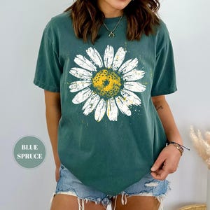 Puede incluir: Una camiseta de color verde azulado con un gran gr&aacute;fico de margarita. La margarita tiene p&eacute;talos blancos y un centro amarillo con un aspecto desgastado y vintage. La camiseta tiene mangas cortas y un corte relajado. El texto "BLUE SPRUCE" est&aacute; en un c&iacute;rculo en la parte inferior izquierda.