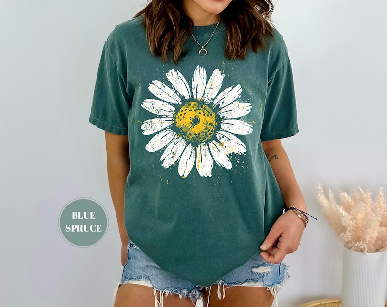 Puede incluir: Una camiseta de color verde azulado con un gran gr&aacute;fico de margarita. La margarita tiene p&eacute;talos blancos y un centro amarillo con un aspecto desgastado y vintage. La camiseta tiene mangas cortas y un corte relajado. El texto "BLUE SPRUCE" est&aacute; en un c&iacute;rculo en la parte inferior izquierda.