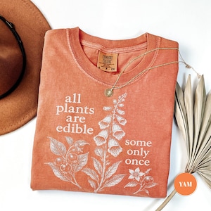 以下が含まれることがあります： テラコッタカラーの半袖Tシャツ。植物のイラストと「all plants are edible some only once」というテキストが白でプリントされています。クルーネックで、襟の内側にComfort Colorsのブランドラベルが付いています。