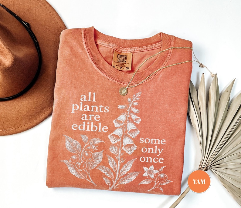 以下が含まれることがあります： テラコッタカラーの半袖Tシャツ。植物のイラストと「all plants are edible some only once」というテキストが白でプリントされています。クルーネックで、襟の内側にComfort Colorsのブランドラベルが付いています。