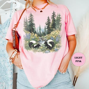 May include: Light pink t-shirt featuring a forest scene with two skunks. The graphic includes a stream, trees, and wildflowers. The shirt has short sleeves and a round neckline. The text "LIGHT PINK" is in a pink circle.