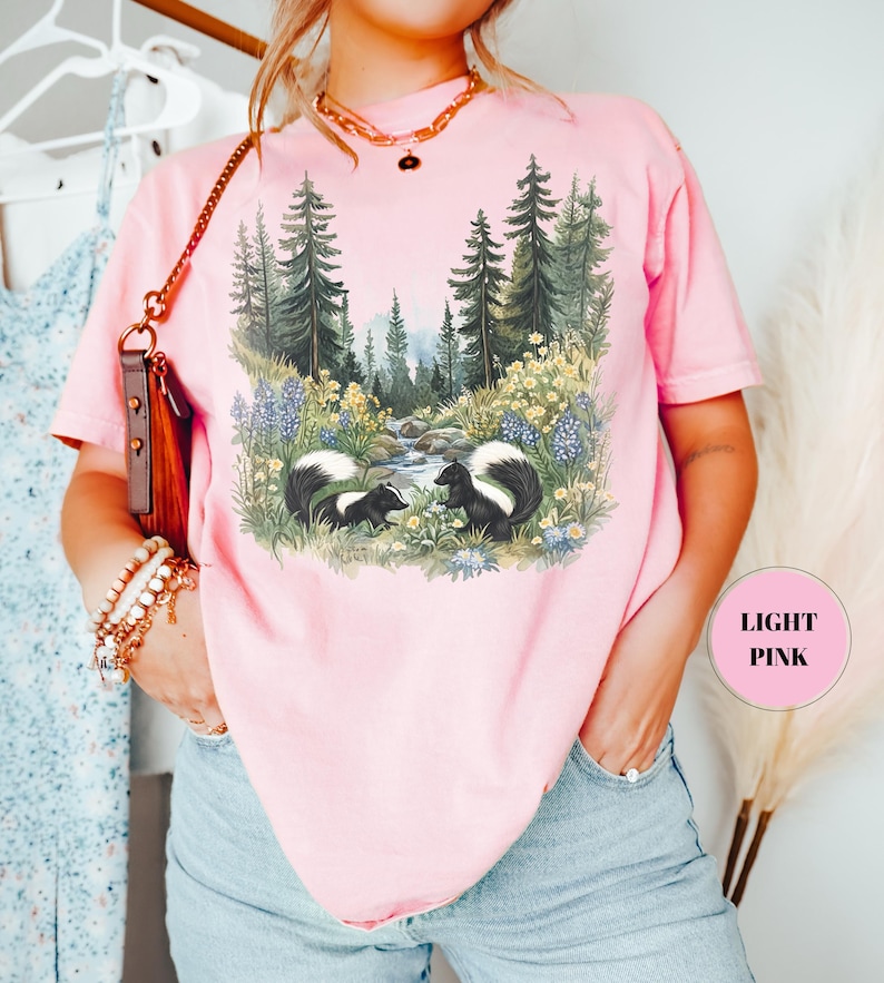 May include: Light pink t-shirt featuring a forest scene with two skunks. The graphic includes a stream, trees, and wildflowers. The shirt has short sleeves and a round neckline. The text "LIGHT PINK" is in a pink circle.