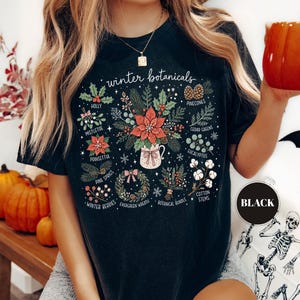 May include: A black t-shirt featuring a graphic print titled winter botanicals. The design displays various illustrations including holly, mistletoe, poinsettia, pinecones, cedar greens, eucalyptus, pine sprigs, winter berries, a wreath, and cotton stems.