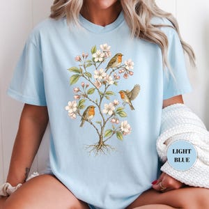 May include: Light blue t-shirt featuring a botanical illustration of a flowering tree with small birds. The design includes white blossoms, green leaves, and three birds with orange and brown feathers. The text "LIGHT BLUE" is in a circle.