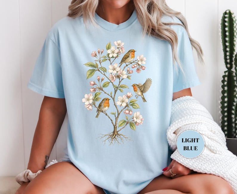 May include: Light blue t-shirt featuring a botanical illustration of a flowering tree with small birds. The design includes white blossoms, green leaves, and three birds with orange and brown feathers. The text "LIGHT BLUE" is in a circle.