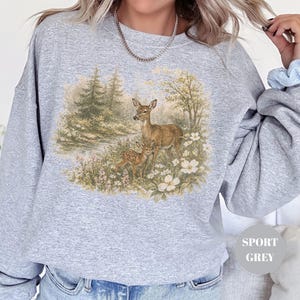 May include: Sport grey sweatshirt featuring a detailed illustration of a doe and fawn in a meadow with white flowers and trees. The design is in a vintage style. The sweatshirt is a comfortable, casual style.