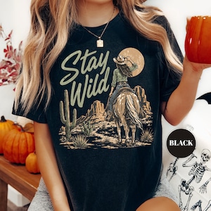 May include: Black t-shirt with a desert-themed graphic. The design features a cowgirl on horseback, cacti, and the words "Stay Wild" in a stylized font. A black circle with the word "BLACK" is in the lower right corner.