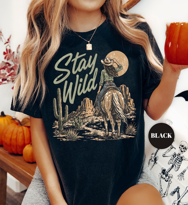 May include: Black t-shirt with a desert-themed graphic. The design features a cowgirl on horseback, cacti, and the words "Stay Wild" in a stylized font. A black circle with the word "BLACK" is in the lower right corner.