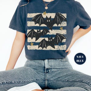 May include: A navy blue short-sleeve t-shirt featuring a graphic print of three black bats flying over horizontal distressed beige stripes. The design includes small gold-colored stars scattered among the bats. A circular navy blue badge reads NAVY BLUE.