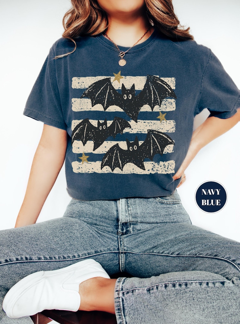 May include: A navy blue short-sleeve t-shirt featuring a graphic print of three black bats flying over horizontal distressed beige stripes. The design includes small gold-colored stars scattered among the bats. A circular navy blue badge reads NAVY BLUE.