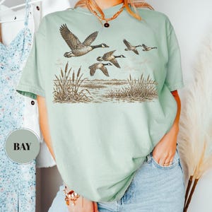 Peut inclure: Un t-shirt &agrave; manches courtes vert sauge clair avec une impression graphique marron et noire repr&eacute;sentant un vol d'oies au-dessus d'un marais avec des roseaux. Le t-shirt a un col rond et une coupe d&eacute;contract&eacute;e. Une &eacute;tiquette ronde indique BAY.