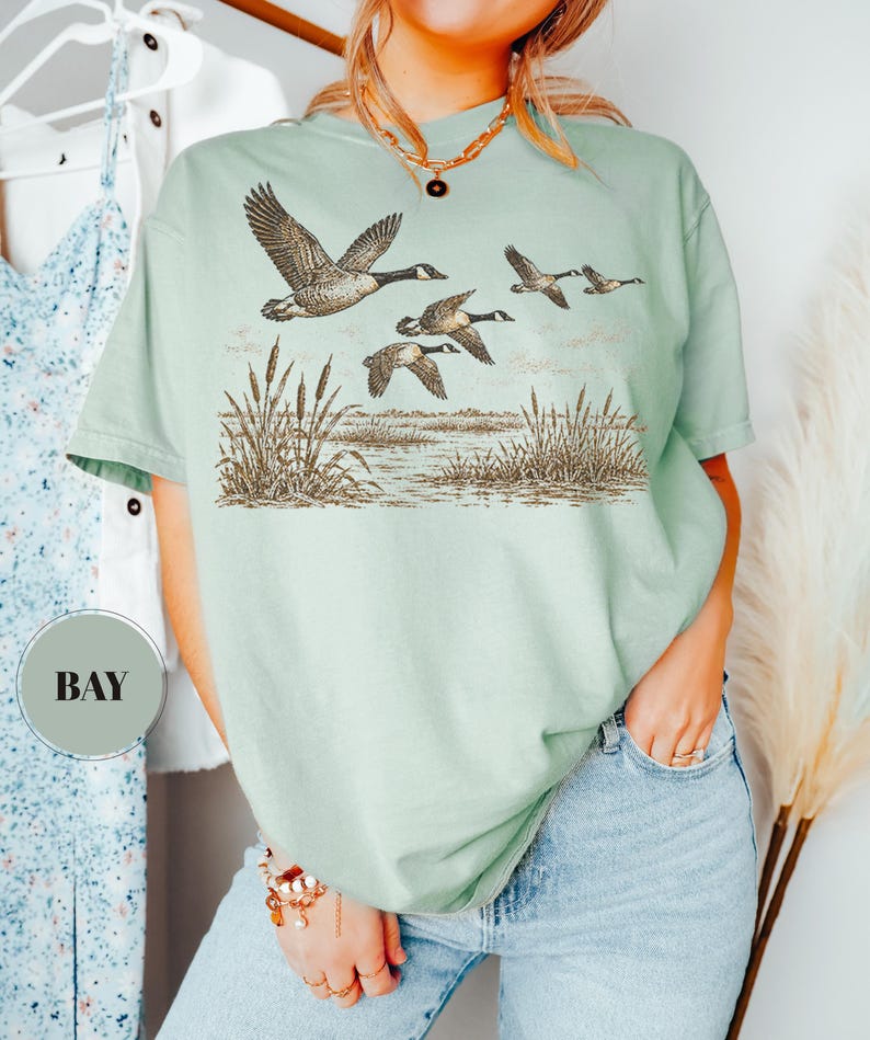 Peut inclure: Un t-shirt &agrave; manches courtes vert sauge clair avec une impression graphique marron et noire repr&eacute;sentant un vol d'oies au-dessus d'un marais avec des roseaux. Le t-shirt a un col rond et une coupe d&eacute;contract&eacute;e. Une &eacute;tiquette ronde indique BAY.