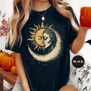 May include: Black t-shirt featuring a celestial design with a sun, moon, and a woman's face. The graphic is in shades of yellow and cream. The shirt has short sleeves and a crew neck.