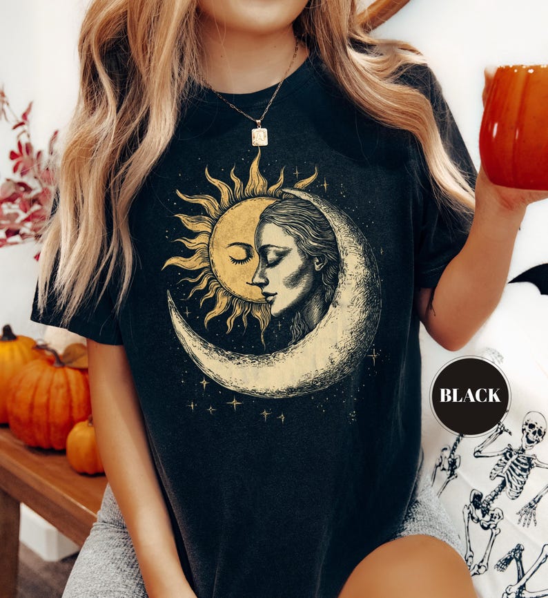 May include: Black t-shirt featuring a celestial design with a sun, moon, and a woman's face. The graphic is in shades of yellow and cream. The shirt has short sleeves and a crew neck.