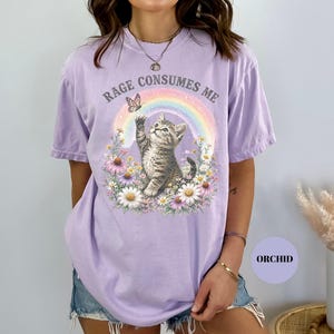 May include: A light purple t-shirt with the text "RAGE CONSUMES ME" above a graphic of a kitten reaching for a butterfly near a rainbow. The kitten is surrounded by flowers.