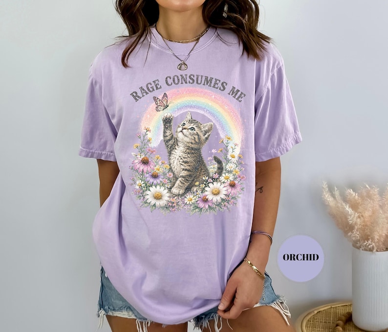 May include: A light purple t-shirt with the text "RAGE CONSUMES ME" above a graphic of a kitten reaching for a butterfly near a rainbow. The kitten is surrounded by flowers.