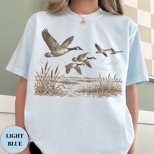 Peut inclure: Un t-shirt bleu clair arborant une illustration d&eacute;taill&eacute;e d'un vol d'oies au-dessus d'un marais avec des massettes. Le dessin utilise des tons bruns et beiges pour repr&eacute;senter les oiseaux en plein vol et la v&eacute;g&eacute;tation mar&eacute;cageuse.