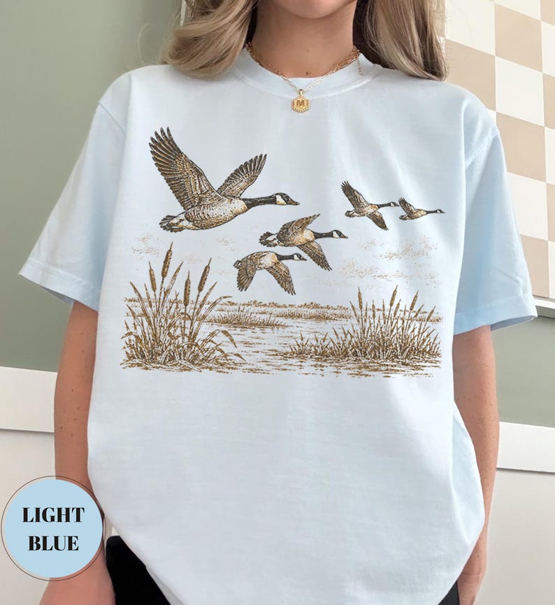 Peut inclure: Un t-shirt bleu clair arborant une illustration d&eacute;taill&eacute;e d'un vol d'oies au-dessus d'un marais avec des massettes. Le dessin utilise des tons bruns et beiges pour repr&eacute;senter les oiseaux en plein vol et la v&eacute;g&eacute;tation mar&eacute;cageuse.