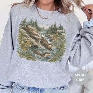 Puede incluir: Una sudadera gris claro de cuello redondo con un estampado gr&aacute;fico detallado de tres nutrias nadando y jugando en un arroyo forestal rodeado de pinos, helechos y flores silvestres amarillas. El texto SPORT GREY es visible en un c&iacute;rculo.