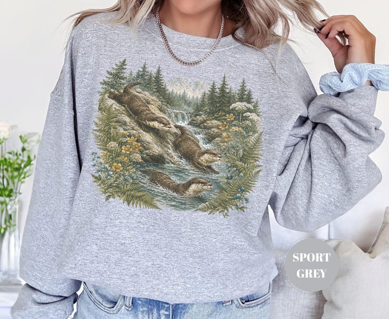 Puede incluir: Una sudadera gris claro de cuello redondo con un estampado gr&aacute;fico detallado de tres nutrias nadando y jugando en un arroyo forestal rodeado de pinos, helechos y flores silvestres amarillas. El texto SPORT GREY es visible en un c&iacute;rculo.
