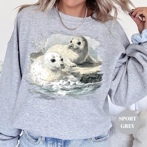 Pode incluir: Camisola cinza desportiva com uma ilustra&ccedil;&atilde;o vintage de duas focas num ambiente marinho. As focas s&atilde;o retratadas em tons de branco e cinza, com detalhes da &aacute;gua e das rochas. A camisola tem um corte folgado.