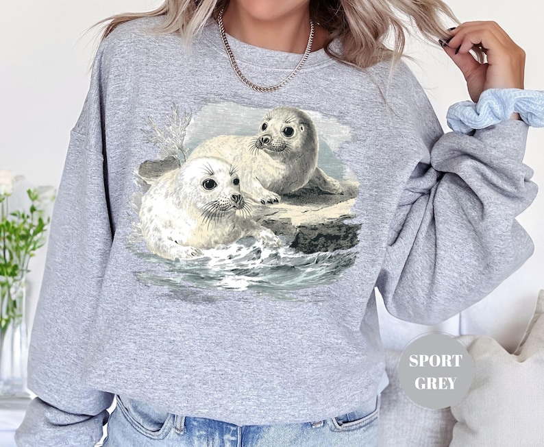 Pode incluir: Camisola cinza desportiva com uma ilustra&ccedil;&atilde;o vintage de duas focas num ambiente marinho. As focas s&atilde;o retratadas em tons de branco e cinza, com detalhes da &aacute;gua e das rochas. A camisola tem um corte folgado.