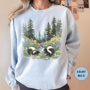 May include: Light blue sweatshirt featuring a nature scene with two skunks near a stream, surrounded by trees and flowers. The sweatshirt has a crew neck and long sleeves. The text "LIGHT BLUE" is in a circle on the bottom right.