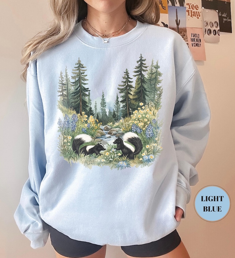 May include: Light blue sweatshirt featuring a nature scene with two skunks near a stream, surrounded by trees and flowers. The sweatshirt has a crew neck and long sleeves. The text "LIGHT BLUE" is in a circle on the bottom right.