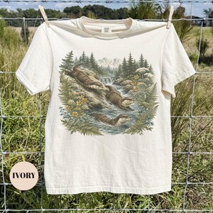 Puede incluir: Una camiseta de manga corta color marfil con un estampado gr&aacute;fico detallado de tres nutrias marrones nadando y jugando en un arroyo rocoso rodeado de pinos, helechos y flores silvestres amarillas con un paisaje monta&ntilde;oso de fondo.