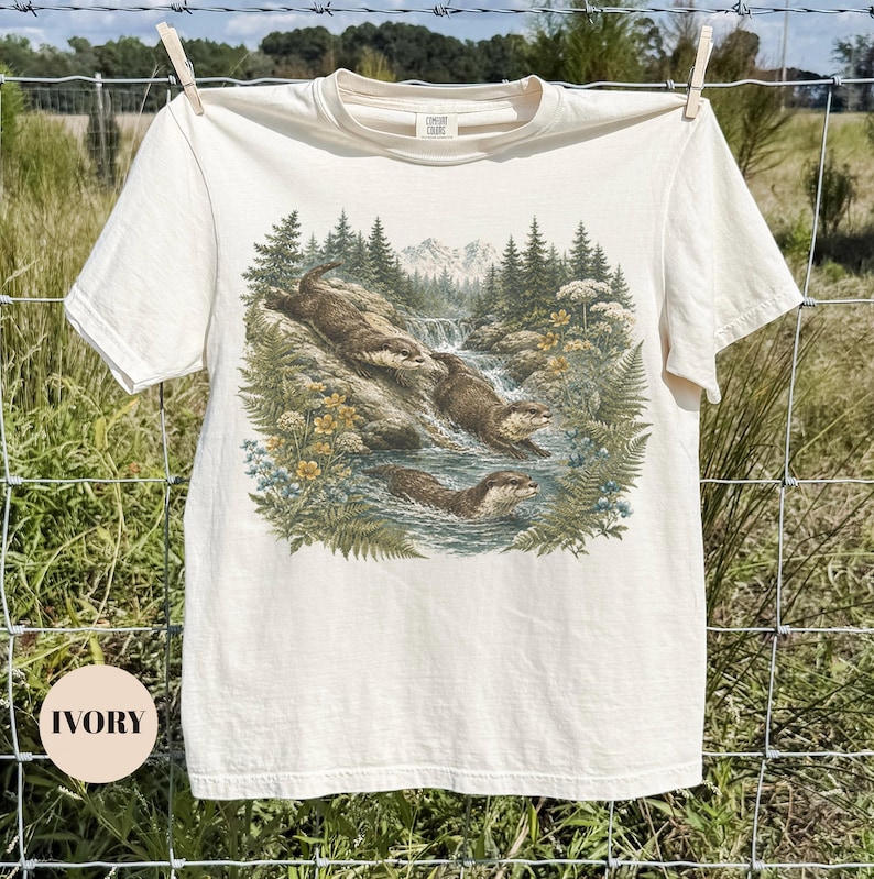 Puede incluir: Una camiseta de manga corta color marfil con un estampado gr&aacute;fico detallado de tres nutrias marrones nadando y jugando en un arroyo rocoso rodeado de pinos, helechos y flores silvestres amarillas con un paisaje monta&ntilde;oso de fondo.