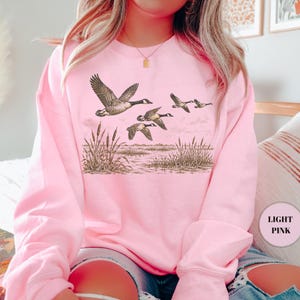 Peut inclure: Un sweat-shirt &agrave; col rond rose clair arborant une impression graphique d&eacute;taill&eacute;e en brun et noir repr&eacute;sentant un vol d'oies au-dessus d'un paysage mar&eacute;cageux avec de grands roseaux. Le motif est centr&eacute; sur la poitrine du v&ecirc;tement.
