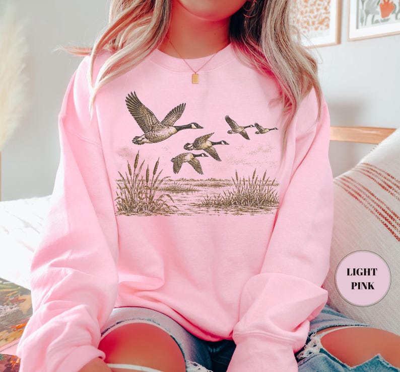 Peut inclure: Un sweat-shirt &agrave; col rond rose clair arborant une impression graphique d&eacute;taill&eacute;e en brun et noir repr&eacute;sentant un vol d'oies au-dessus d'un paysage mar&eacute;cageux avec de grands roseaux. Le motif est centr&eacute; sur la poitrine du v&ecirc;tement.
