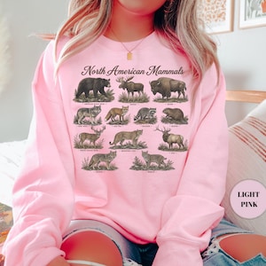 May include: A light pink crewneck sweatshirt featuring the text North American Mammals and detailed illustrations of various animals including a black bear, moose, bison, gray wolf, red fox, raccoon, beaver, white-tailed deer, mountain lion, elk, coyote, and bobcat.