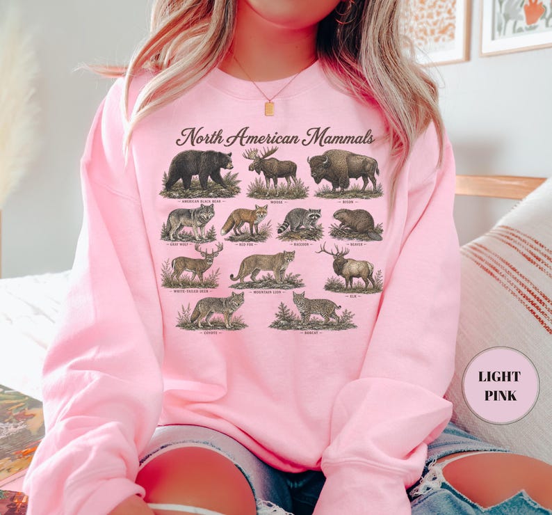 May include: A light pink crewneck sweatshirt featuring the text North American Mammals and detailed illustrations of various animals including a black bear, moose, bison, gray wolf, red fox, raccoon, beaver, white-tailed deer, mountain lion, elk, coyote, and bobcat.