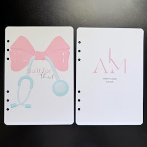 May include: Two white planner inserts. One features a pink bow and a stethoscope with the text "Built for this!". The other has the letters "AM" and the text "Indestructible but soft."