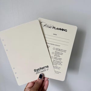 May include: Two off-white planner inserts with the text "Wish Planning" and "Systems Thinker". One insert has a checklist and the other has lines for notes. The inserts are held against a white background.