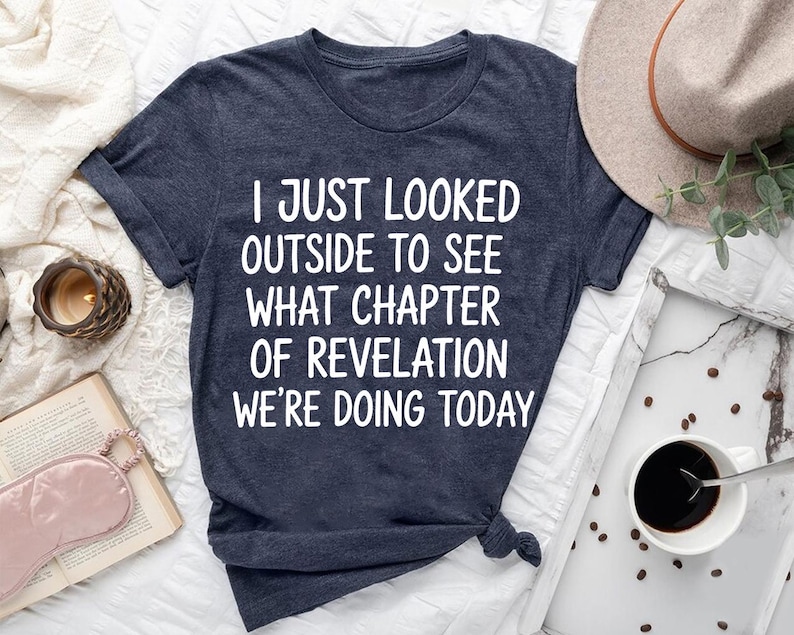 a dark blue t-shirt with white text, a hat, a cup of coffee, a book, and some other items arranged on a bed or a table.