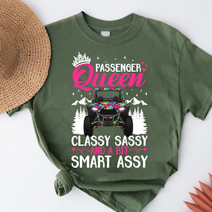 May include: Olive green t-shirt with a graphic that reads "Passenger Queen" above a colorful off-road vehicle design. The text "Classy Sassy And A Bit Smart Assy" is below the vehicle. A straw hat is visible in the upper left corner.