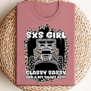 May include: A mauve crew neck t-shirt featuring a graphic of a side-by-side off-road vehicle over a leopard print background. The text reads SXS GIRL CLASSY SASSY AND A BIT SMART ASSY in white lettering with black outlines centered on the front.